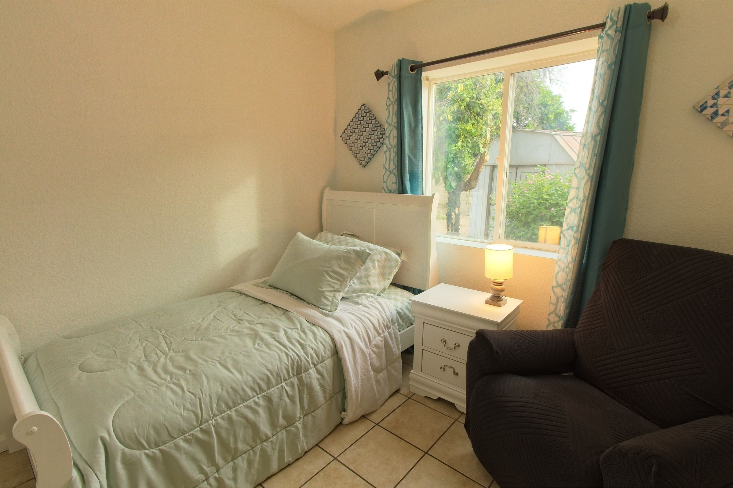 A private bedroom with a made single bed, a white nightstand and lamp, a recliner and a large window